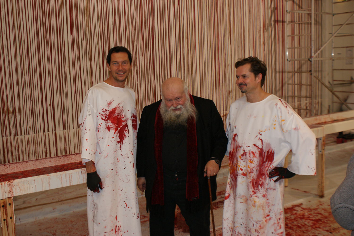 Hermann Nitsch mit zwei Männern vor einer Wand aus roten Farblinien.